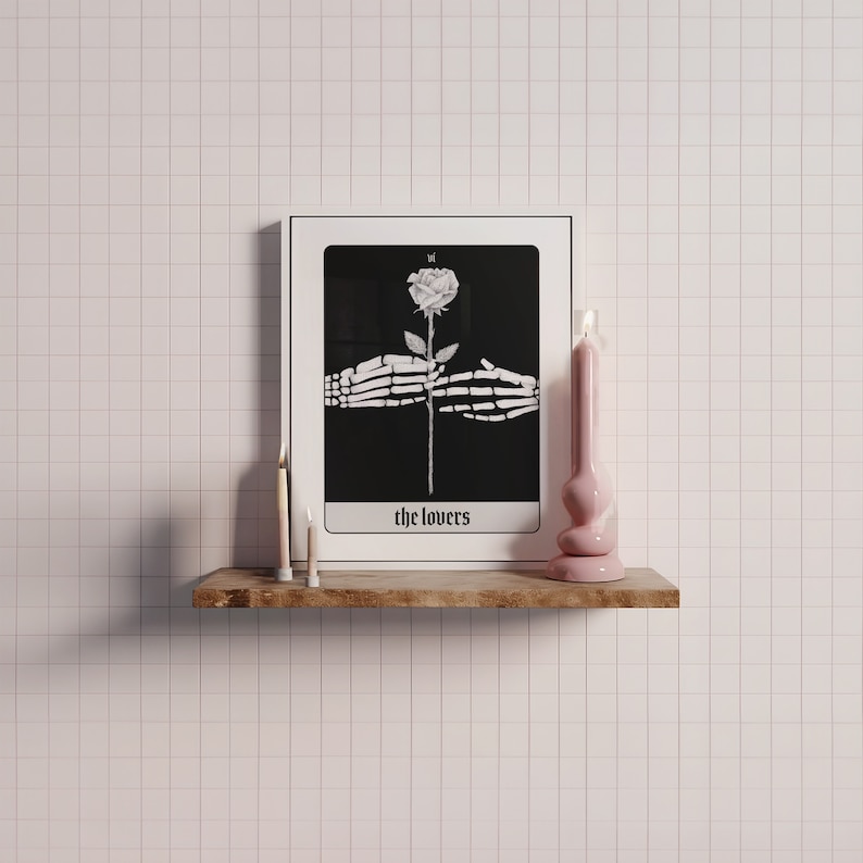 Puede incluir: Una impresi&oacute;n de carta de tarot en blanco y negro con una mano esquel&eacute;tica sosteniendo una rosa blanca. La carta se titula "The Lovers" y est&aacute; enmarcada en un marco blanco. La impresi&oacute;n se muestra en un estante de madera con dos velas blancas y una vela rosa.