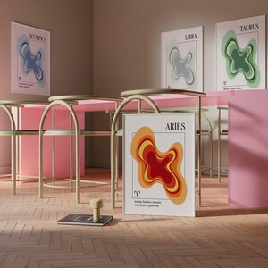 May include: A room setting featuring four framed zodiac art prints: Scorpio, Libra, Taurus, and Aries. Each print displays a layered, abstract design in various colors. The Aries print is leaning against a pink wall. The room has pink tables and gold chairs.