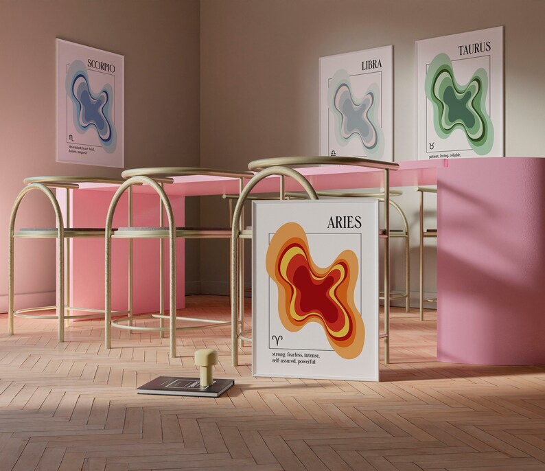 May include: A room setting featuring four framed zodiac art prints: Scorpio, Libra, Taurus, and Aries. Each print displays a layered, abstract design in various colors. The Aries print is leaning against a pink wall. The room has pink tables and gold chairs.