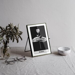 Puede incluir: Una carta de tarot en blanco y negro con una mano esquel&eacute;tica sosteniendo una rosa blanca. La carta se titula "Los Amantes" y se muestra en un marco de metal negro.