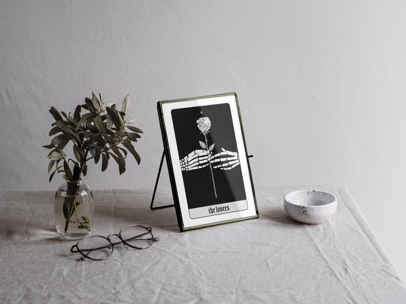 Puede incluir: Una carta de tarot en blanco y negro con una mano esquel&eacute;tica sosteniendo una rosa blanca. La carta se titula "Los Amantes" y se muestra en un marco de metal negro.