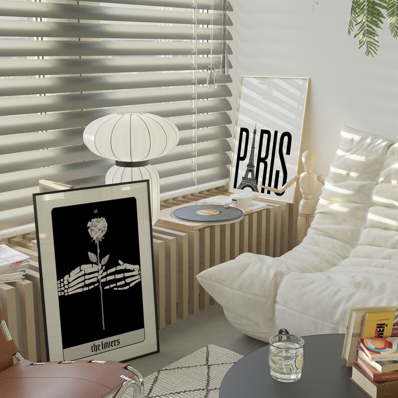 Puede incluir: Una ilustraci&oacute;n en blanco y negro de una mano esquel&eacute;tica sosteniendo una rosa con el texto "the lovers" debajo. La ilustraci&oacute;n est&aacute; enmarcada y colgada en una pared. Una l&aacute;mpara blanca con una pantalla blanca est&aacute; sobre una mesa frente al marco. Un tocadiscos est&aacute; sobre una mesa en el fondo. La pared detr&aacute;s del marco es blanca y tiene una ventana con persianas. La ventana est&aacute; abierta y las persianas est&aacute;n cerradas. Hay un sof&aacute; blanco en el fondo. El sof&aacute; est&aacute; hecho de tela blanca y tiene una almohada blanca. El sof&aacute; est&aacute; frente a la ventana. La habitaci&oacute;n est&aacute; decorada en un estilo moderno.