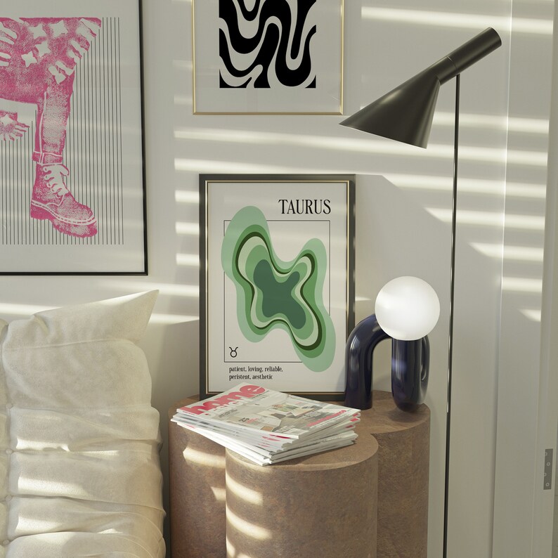 May include: A modern interior scene featuring framed art, a floor lamp, and a decorative table. The artwork includes a Taurus zodiac print and abstract designs. A small, dark blue lamp with a white globe sits on the table, next to a stack of magazines.