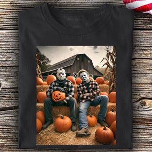 May include: A black t-shirt featuring a graphic print of two figures wearing horror movie masks, one with a hockey mask and one with a white mask, sitting on hay bales in front of a barn surrounded by orange pumpkins in a field setting.