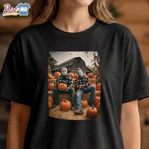 May include: A black t-shirt featuring a graphic print of two figures wearing horror masks, one in a plaid shirt and one in a flannel shirt, sitting on hay bales surrounded by orange pumpkins in front of a dark wooden barn structure.