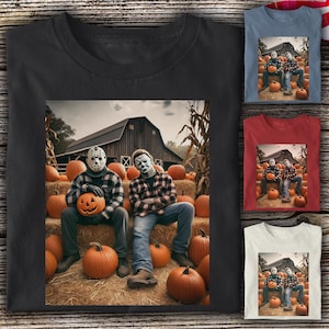 May include: A black t-shirt featuring a graphic of two figures wearing horror masks, one in a plaid shirt and one in a flannel shirt, sitting on hay bales in front of a barn with pumpkins. The design is also shown on blue, red, and white shirts.