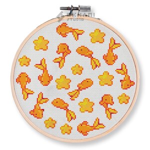 Puede incluir: Un aro de bordado blanco con un diseño de punto de cruz de peces dorados naranjas y estrellas amarillas sobre un fondo blanco.