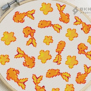 K&ouml;nnte beinhalten: Ein wei&szlig;er Stickrahmen mit einem Kreuzstich-Design aus orangefarbenen Goldfischen und gelben Blumen. Das Design ist auf einem wei&szlig;en Stoffhintergrund.