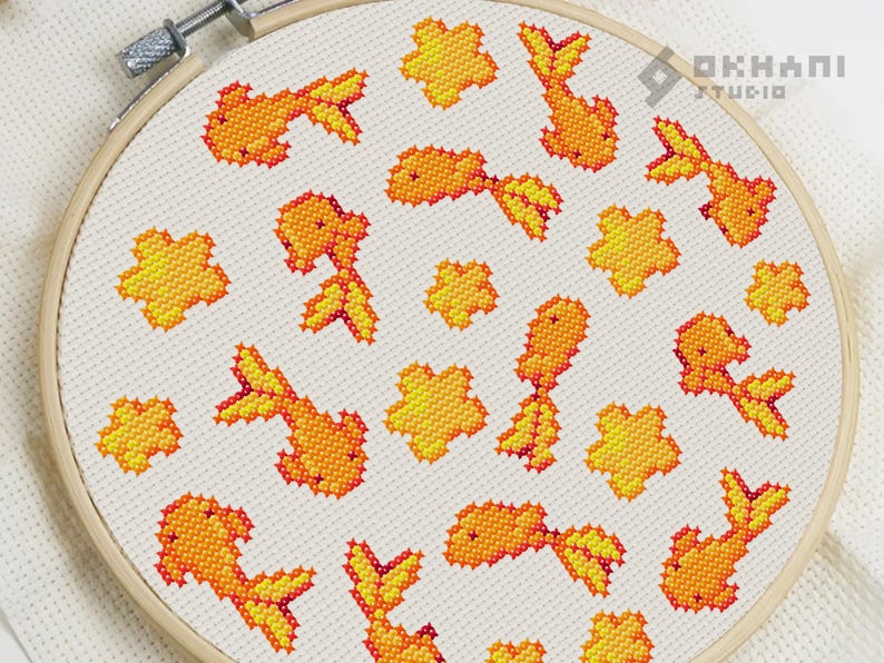 K&ouml;nnte beinhalten: Ein wei&szlig;er Stickrahmen mit einem Kreuzstich-Design aus orangefarbenen Goldfischen und gelben Blumen. Das Design ist auf einem wei&szlig;en Stoffhintergrund.