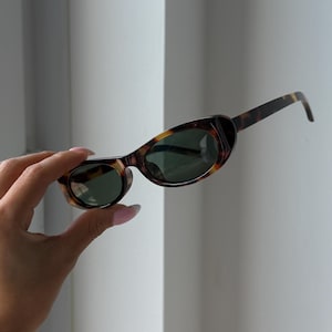 May include: Oval-shaped sunglasses with a tortoiseshell pattern frame and dark green lenses. The sunglasses are held up against a white background. The arms of the sunglasses are thin and have a matching tortoiseshell pattern.