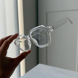 Może przedstawiać: Przezroczyste okulary w kwadratowych oprawkach. Okulary mają przezroczystą oprawkę i soczewki. Zausszniki są również przezroczyste i lekko zakrzywione. Okulary są trzymane na białym tle.