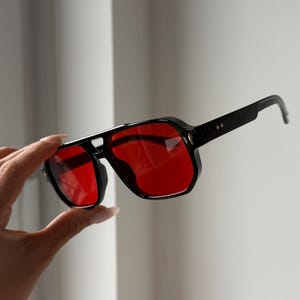 May include: Black sunglasses with a square frame and red lenses. The sunglasses have a thick, angular design with a bridge connecting the lenses. The temples are thin and extend to the ear pieces. The sunglasses are held up by a hand.