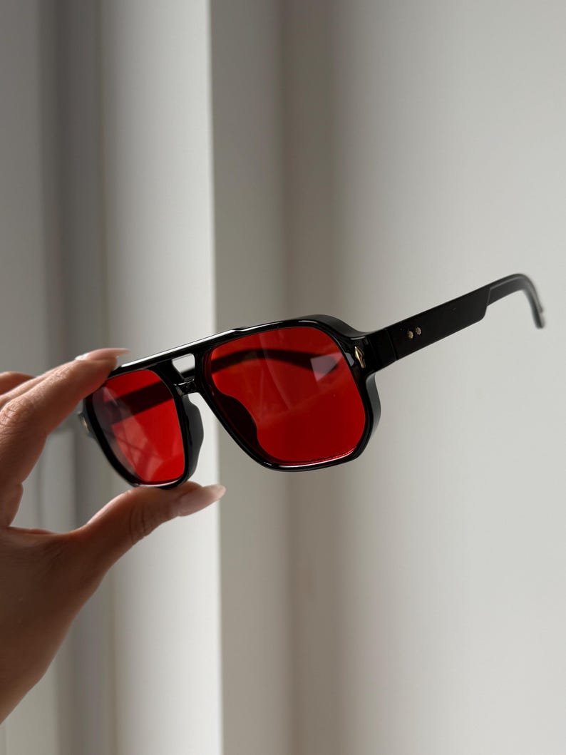 May include: Black sunglasses with a square frame and red lenses. The sunglasses have a thick, angular design with a bridge connecting the lenses. The temples are thin and extend to the ear pieces. The sunglasses are held up by a hand.
