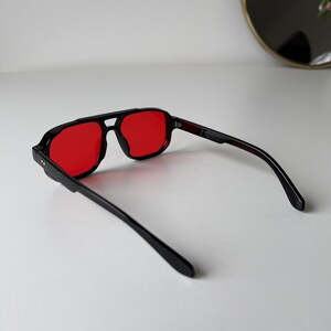 May include: Black-framed sunglasses with red lenses. The sunglasses have a square shape and a double bridge. The temples are black and extend back from the frame. The sunglasses are on a white surface.