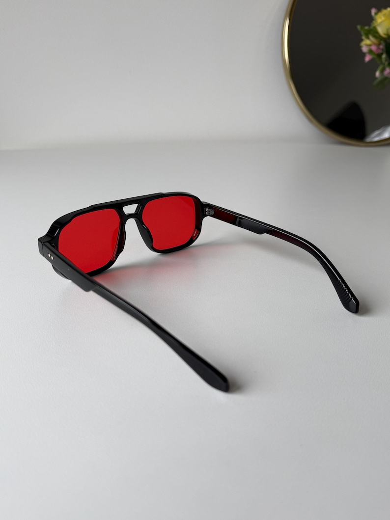 May include: Black-framed sunglasses with red lenses. The sunglasses have a square shape and a double bridge. The temples are black and extend back from the frame. The sunglasses are on a white surface.