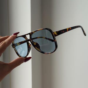 May include: Tortoiseshell-patterned sunglasses with blue-tinted lenses. The sunglasses have a classic aviator shape with a double bridge and thick frames. The arms of the sunglasses are also tortoiseshell, and the lenses are a light blue color.