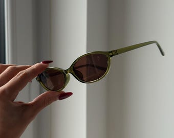 Könnte beinhalten: Olivgrüne ovale Sonnenbrille mit braunen Gläsern. Die Sonnenbrille hat einen dünnen Rahmen und Bügel. Die Gläser sind dunkelbraun. Die Sonnenbrille wird von einer Hand mit rotem Nagellack gehalten.