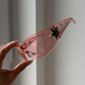 May include: Pink sunglasses with a star design. The sunglasses have a light pink frame and lenses. Silver stars are placed on the top of the lenses. The sunglasses are held by a hand.