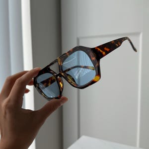 Puede incluir: Gafas de sol carey con lentes azules. Las gafas de sol tienen una forma geométrica y una montura gruesa. Las patillas de las gafas están curvadas. Las lentes son de color azul claro.