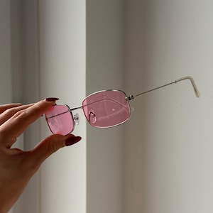 May include: Silver-framed sunglasses with rectangular pink lenses. The glasses have a thin metal frame and temples. The lenses are a vibrant pink color, and the overall design is sleek and modern. The sunglasses are held up against a white background.