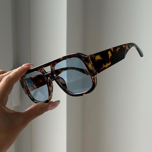 May include: Tortoiseshell sunglasses with blue lenses. The sunglasses have a thick frame and a double bridge design. The lenses are a light blue color, and the frame has a brown and black pattern.