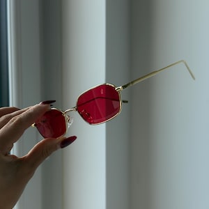 Puede incluir: Gafas de sol con montura dorada y lentes rojas. Las gafas tienen forma rectangular y están sujetas por una mano con uñas largas de color rojo oscuro. El fondo es de color gris claro.