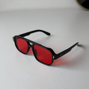 May include: Black aviator sunglasses with red lenses. The sunglasses feature a thick black frame, a double bridge, and straight temples. The lenses are a bright red, and the design is modern and stylish, suitable for various occasions.