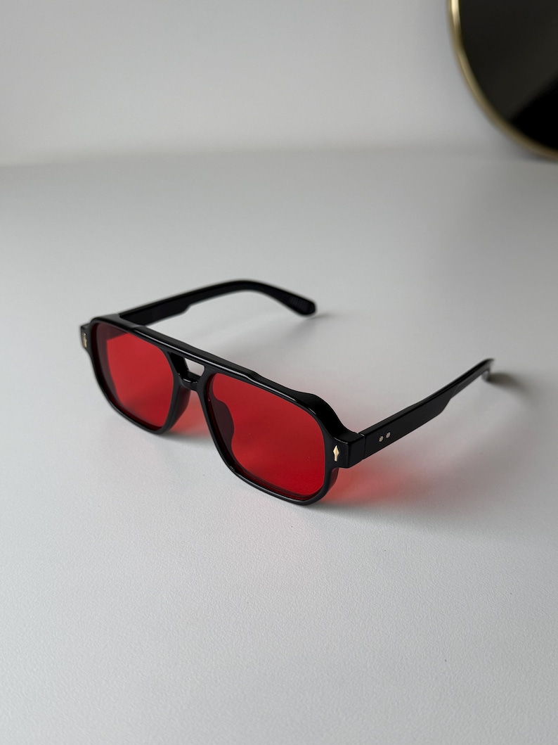 May include: Black aviator sunglasses with red lenses. The sunglasses feature a thick black frame, a double bridge, and straight temples. The lenses are a bright red, and the design is modern and stylish, suitable for various occasions.