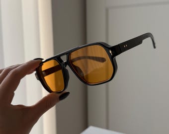 Può includere: Occhiali da sole neri con lenti colorate arancioni. Gli occhiali hanno un design a montatura quadrata con doppio ponte e aste spesse. Le lenti sono color ambra e le montature sono nere lucide.