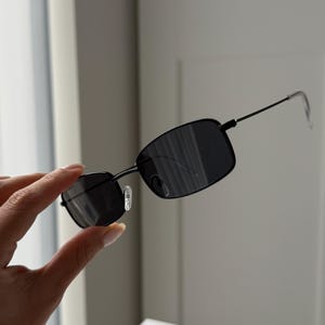 Puede incluir: Gafas de sol rectangulares negras con lentes oscuras y montura fina de metal negro. Las gafas de sol están sujetas por una mano. Las lentes son reflectantes y las patillas son finas.