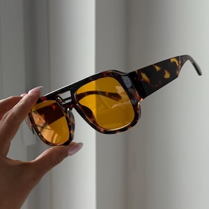 Puede incluir: Gafas de sol cuadradas con estampado de carey y lentes de color amarillo. Las gafas tienen una montura gruesa y un puente que conecta las lentes. Las patillas de las gafas también tienen un estampado. Sobre un fondo blanco.