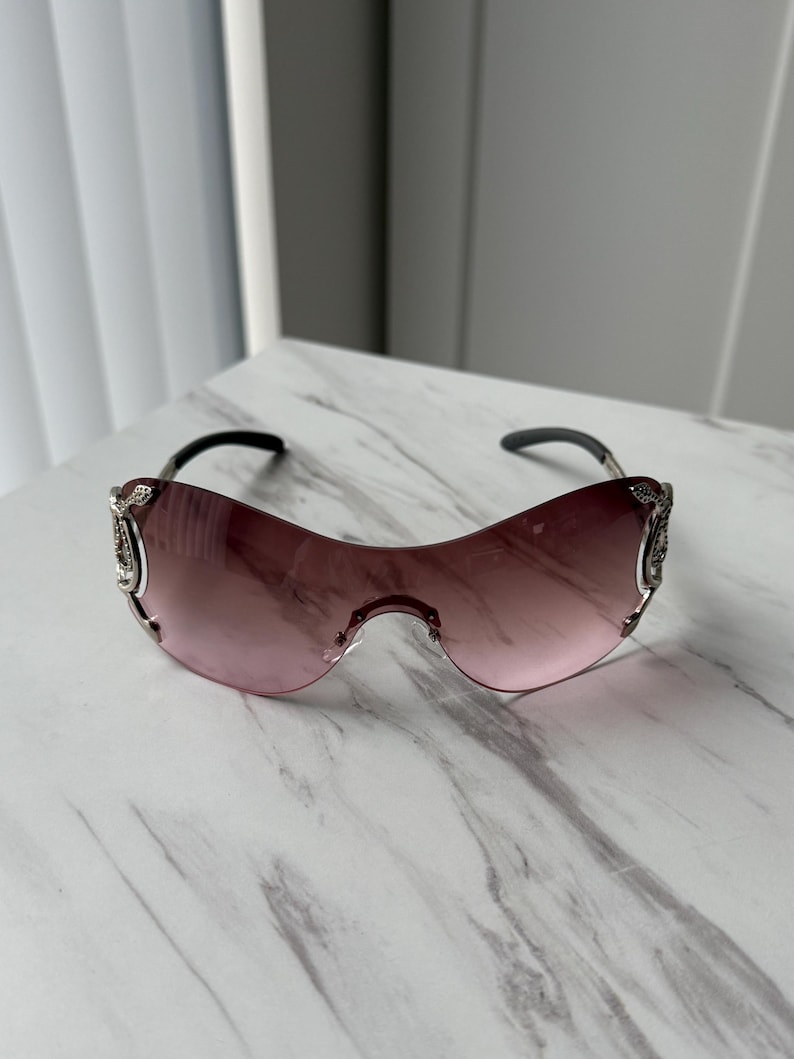 May include: A pair of pink tinted sunglasses with silver accents and a decorative design on the arms.