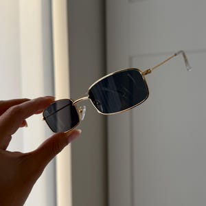 May include: Gold-framed square sunglasses with black lenses. The sunglasses are held in a hand.