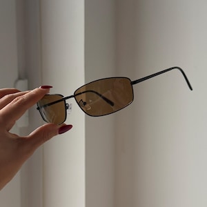 May include: Black-framed sunglasses with brown lenses. The rectangular glasses are held up by a hand. The sunglasses have a thin frame and a classic design. The lenses are tinted brown.