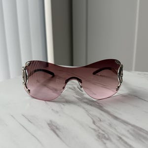May include: A pair of pink tinted sunglasses with silver frames and decorative accents. The sunglasses have a unique, curved shape.