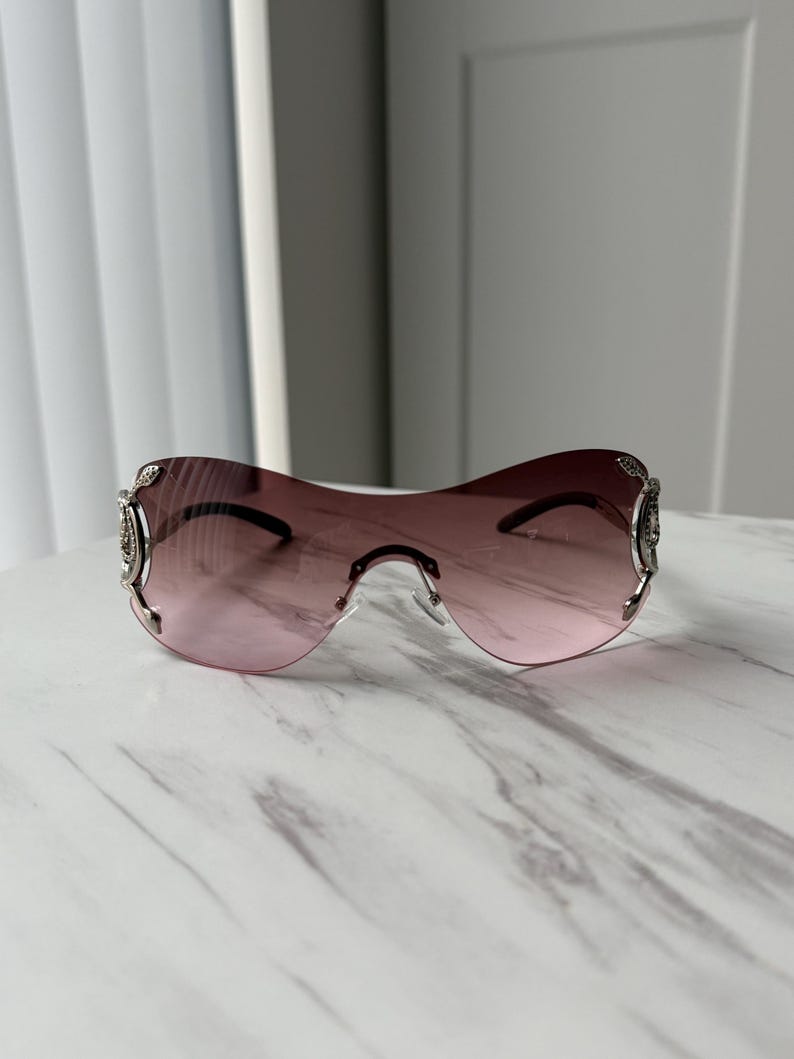 May include: A pair of pink tinted sunglasses with silver frames and decorative accents. The sunglasses have a unique, curved shape.
