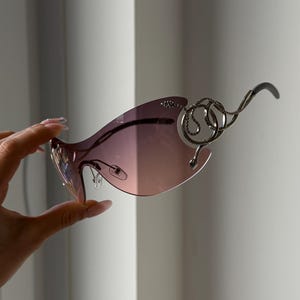May include: Fashionable sunglasses with a pink gradient lens and detailed silver metalwork. The frame has a distinctive, curved shape with decorative elements near the temples. The sunglasses are held up by a hand.