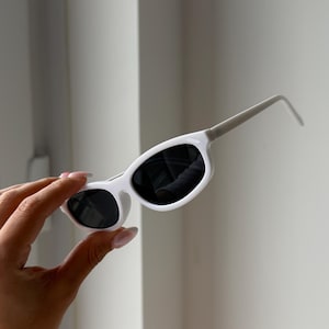 May include: White sunglasses with black lenses. The sunglasses have a sleek, modern design with a thick frame and thin arms. The sunglasses are held up by a hand against a white background.