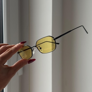 May include: Black rectangular sunglasses with yellow lenses. The sunglasses have a thin black frame and are held up by a hand with red nail polish. The lenses are a bright yellow color.