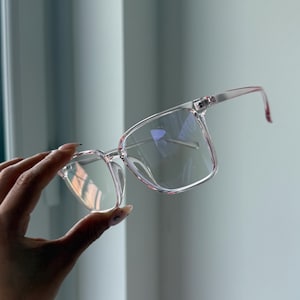 Könnte beinhalten: Brille mit transparentem Rahmen und rosa Farbton. Die Brille hat eine quadratische Form und wird von einer Hand gehalten. Die Gläser sind klar und der Rahmen ist durchscheinend rosa. Der Hintergrund ist hellgrau.