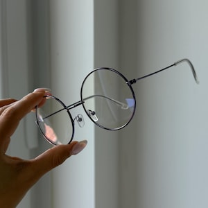 Può includere: Occhiali con montatura rotonda e una sottile montatura in metallo scuro. Le lenti sono trasparenti e gli occhiali sono tenuti da una mano. Il design generale è minimalista e moderno, adatto all'uso quotidiano. Gli occhiali hanno un'estetica classica e intellettuale.