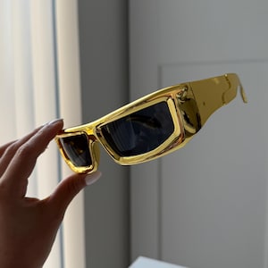 Może przedstawiać: Złote okulary przeciwsłoneczne z ciemnymi soczewkami. Okulary mają grubą, prostokątną oprawkę i elegancki, nowoczesny design. Okulary są trzymane na neutralnym tle.
