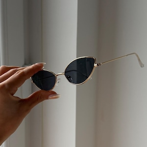Può includere: Occhiali da sole dorati a forma di occhi di gatto con lenti scure. Le sottili montature in metallo dorato hanno un design delicato. Gli occhiali da sole sono tenuti su uno sfondo bianco.