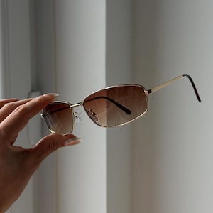Peut inclure: Lunettes de soleil à monture dorée avec verres dégradés bruns. Les montures rectangulaires ont un design classique, et les branches sont fines et dorées. Les verres passent d'une teinte plus claire en haut à un brun plus foncé en bas.
