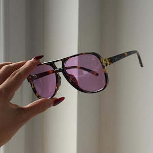 May include: Aviator-style sunglasses with a tortoiseshell frame and purple-tinted lenses. The sunglasses have a double bridge and a classic, retro design. The arms of the sunglasses are also tortoiseshell, providing a cohesive look.
