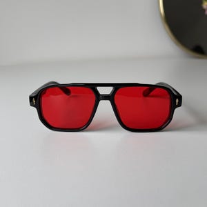 May include: Black sunglasses with red lenses. The sunglasses have a square shape with a double bridge and a small decorative detail on each temple. The frames are thick and the lenses are a vibrant red colour.