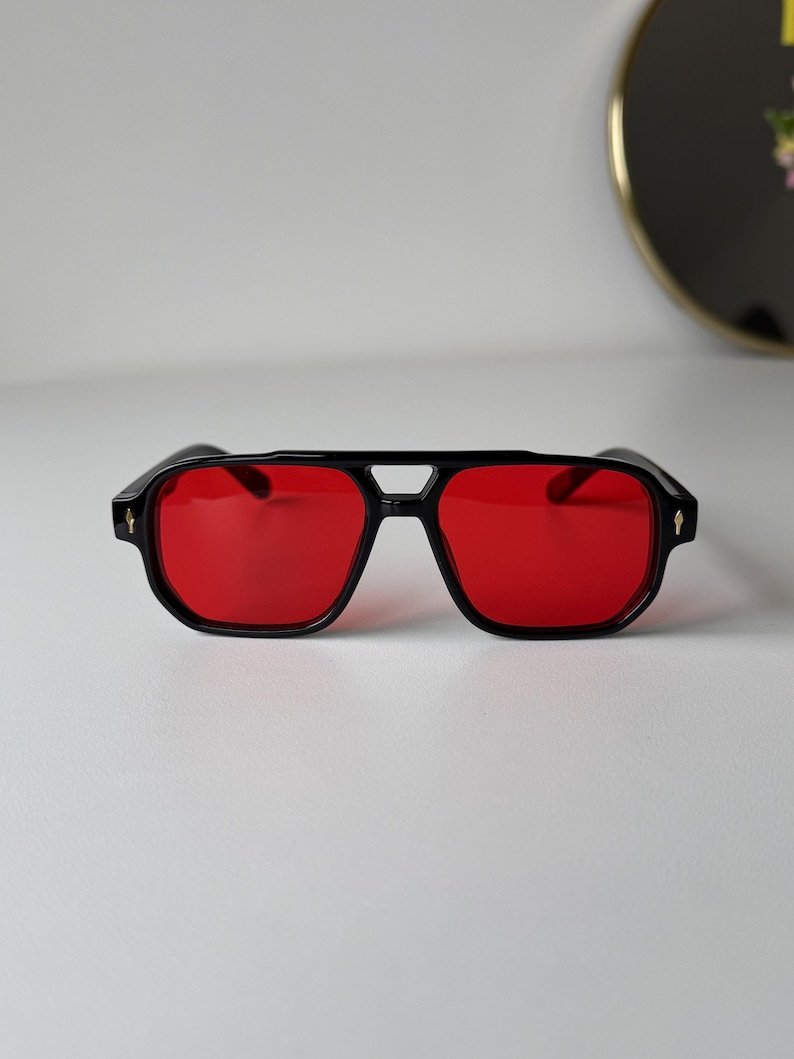 May include: Black sunglasses with red lenses. The sunglasses have a square shape with a double bridge and a small decorative detail on each temple. The frames are thick and the lenses are a vibrant red colour.