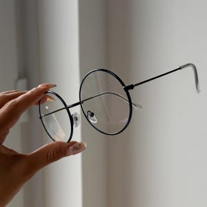 May include: Round, black-rimmed spectacles with clear lenses. The spectacles have a thin, metal frame. The design is simple and classic. The spectacles are held against a neutral background.