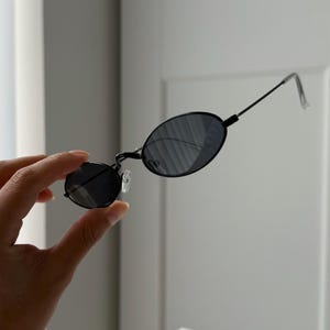 Puede incluir: Gafas de sol ovaladas negras con montura fina negra y lentes oscuras. Las gafas de sol tienen un diseño clásico y retro. Las patillas son finas y se extienden hasta las patillas transparentes.