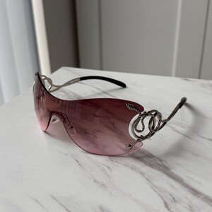 May include: Pink sunglasses with a silver frame and intricate detailing. The frame features a decorative design resembling a snake.